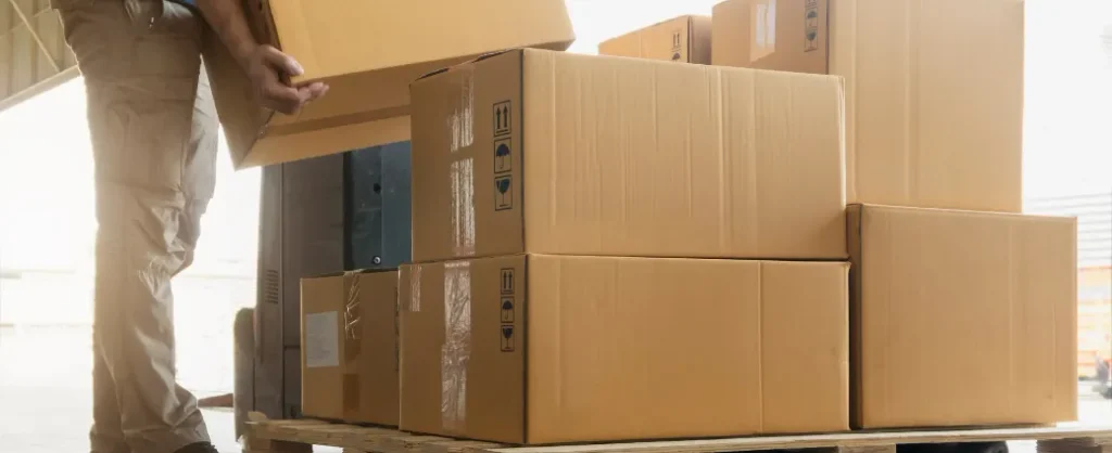 A warehouse worker in beige pants stacking cardboard boxes onto a wooden pallet, preparing them for consolidation and repacking.