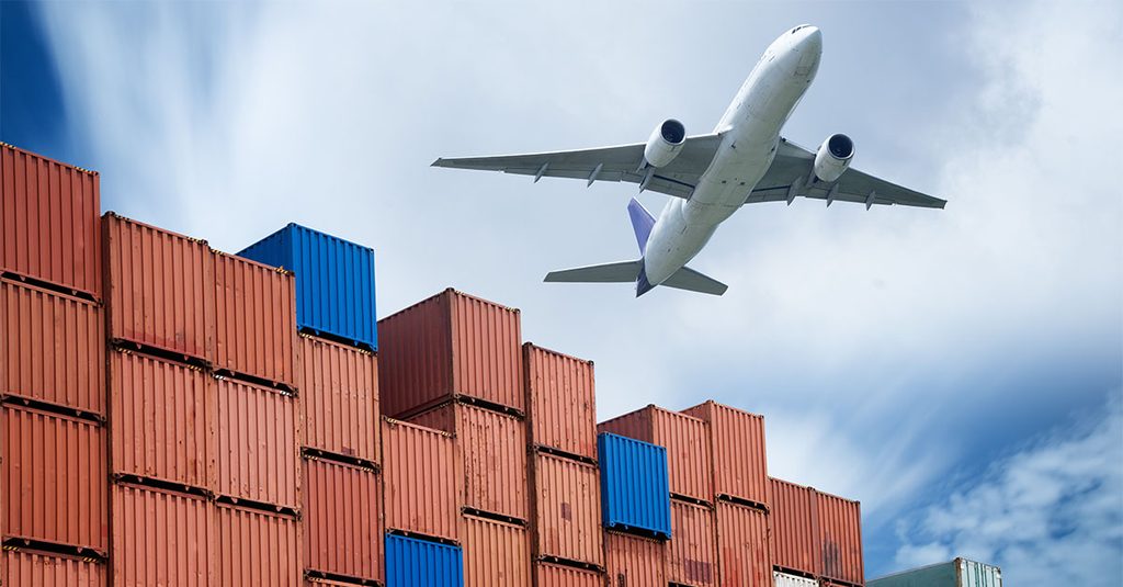 A large cargo airplane flying over towering stacks of red and blue shipping containers under a cloudy blue sky.