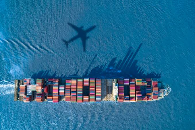 A top-down aerial view of a cargo ship loaded with colorful shipping containers sailing on deep blue water, with the distinct shadow of an airplane cast overhead.